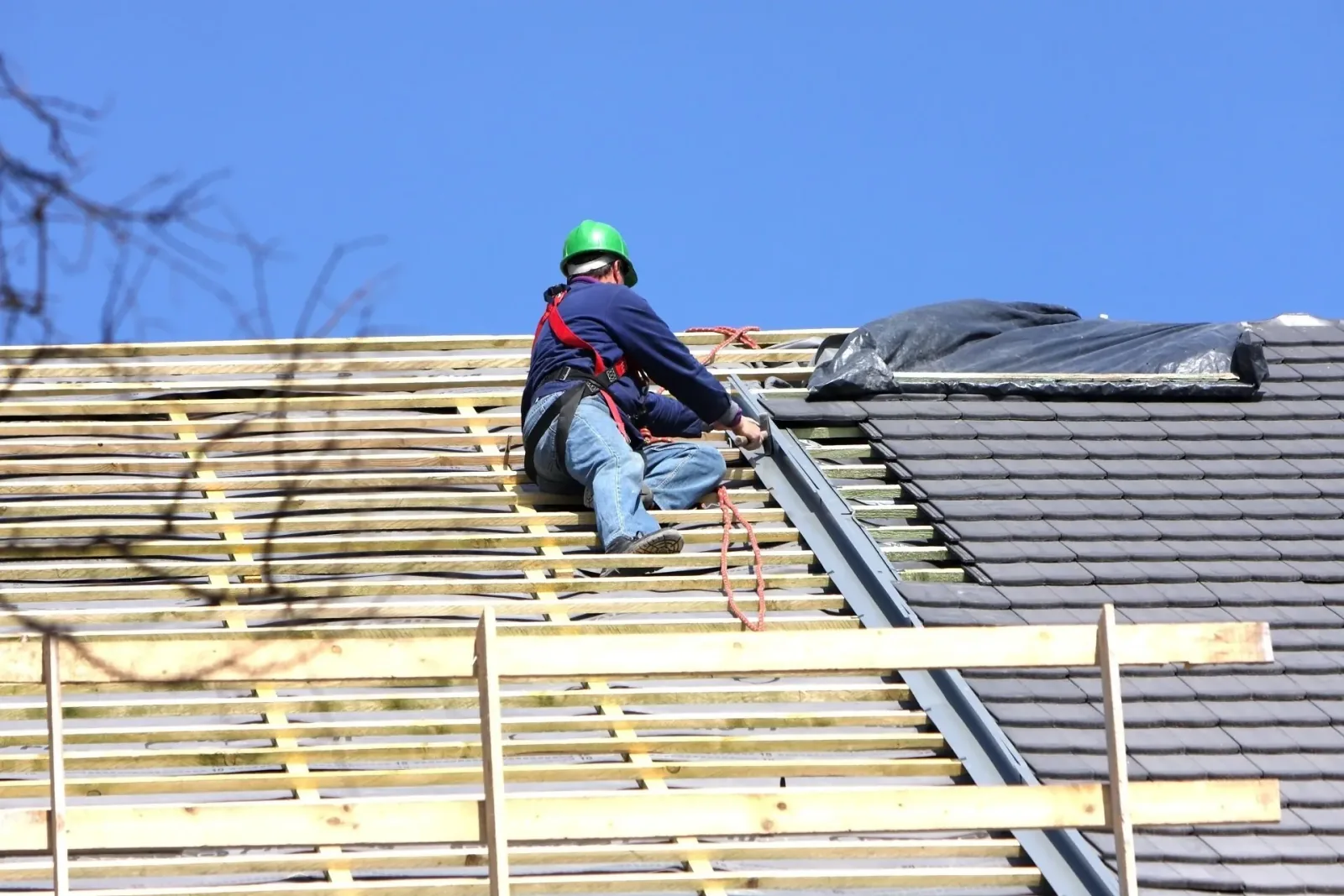 Roofing contractor in a safety harness fitting battens and shingles across a pitched residential roof